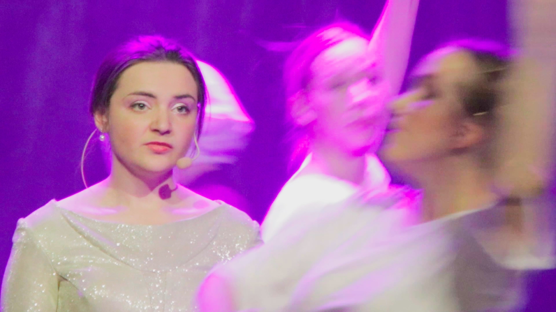 Skådespelerska i glittrande klänning omgiven av dansare i lila scenljus